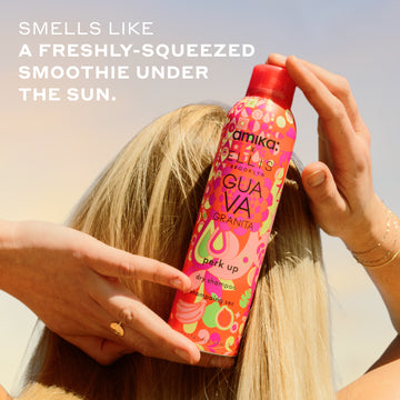 Woman with blonde hair and gold ring and bracelets holds Amika x Ellis Brooklyn GUAVA GRANITA Perk Up Dry Shampoo against the back of her head against blue and yellow sky with white text that reads " smells like a freshly-squeezed smoothie under the sun"