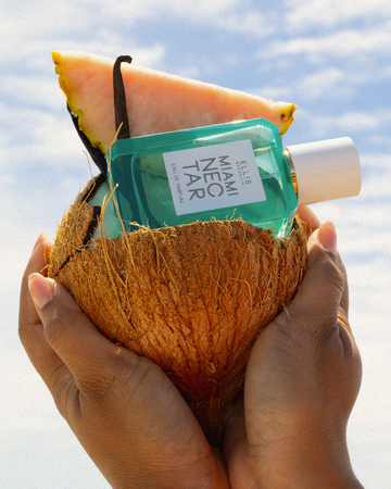 Hands hold a coconut filled with MIAMI NECTAR Eau de Parfum 50ml, a vanilla bean, and a pink pineapple slice against a background of blue sky and white clouds