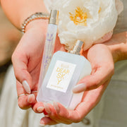 Model with nude nail polish, wearing silver rings, gold and silver bracelets, and white dress, holding Dear Sky 10ml Eau De Parfum Travel Spray, Dear Sky 50ml Eau De Parfum with cap removed, and white flower in both hands.