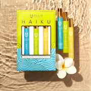 Green, blue, and gold Haiku Fragrance Rollerball Trio Box showing Salt 10ml Fragrance Rollerball, Sun Fruit 10ml Fragrance Rollerball, and Sweet 10ml Fragrance Rollerball, on beige water look backdrop with Salt 10ml Fragrance Rollerball, Sun Fruit 10ml Fragrance Rollerball, Sweet 10ml Fragrance Rollerball, and white flower.
