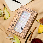 Memoir Fragrance Discovery Set box with beige and black floral design showing Dear Sky Eau De Parfum 1.5ml Vial, Miami Nectar Eau De Parfum 1.5ml Vial, Apple Love Eau De Parfum 1.5ml Vial, Salt Eau De Parfum 1.5ml Vial, Sun Fruit Eau De Parfum 1.5ml Vial, Florist Eau De Parfum 1.5ml Vial, Myth Eau De Parfum 1.5ml Vial, Sweet Eau De Parfum 1.5ml Vial, and Vanilla Milk Eau De Parfum 1.5ml Vial, on tan fabric with fig, pineapple, apple, sandalwood, vanilla bean, pear, white flowers, and notebook with pen.