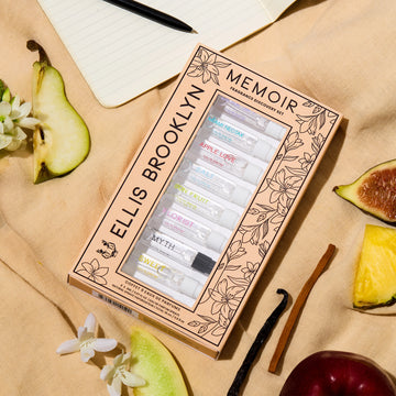 Memoir Fragrance Discovery Set box with beige and black floral design showing Dear Sky Eau De Parfum 1.5ml Vial, Miami Nectar Eau De Parfum 1.5ml Vial, Apple Love Eau De Parfum 1.5ml Vial, Salt Eau De Parfum 1.5ml Vial, Sun Fruit Eau De Parfum 1.5ml Vial, Florist Eau De Parfum 1.5ml Vial, Myth Eau De Parfum 1.5ml Vial, Sweet Eau De Parfum 1.5ml Vial, and Vanilla Milk Eau De Parfum 1.5ml Vial, on tan fabric with fig, pineapple, apple, sandalwood, vanilla bean, pear, white flowers, and notebook with pen.