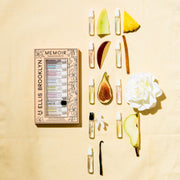 Memoir Fragrance Discovery Set box with beige and black floral design next to Dear Sky Eau De Parfum 1.5ml Vial, Miami Nectar Eau De Parfum 1.5ml Vial, Apple Love Eau De Parfum 1.5ml Vial, Salt Eau De Parfum 1.5ml Vial, Sun Fruit Eau De Parfum 1.5ml Vial, Florist Eau De Parfum 1.5ml Vial, Myth Eau De Parfum 1.5ml Vial, Sweet Eau De Parfum 1.5ml Vial, and Vanilla Milk Eau De Parfum 1.5ml Vial, on yellow background with fig, white flower, honeydew, vanilla bean, sandalwood, pineapple, and apple.
