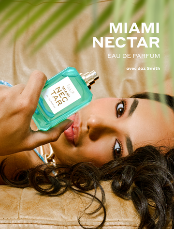 Gros plan sur des feuilles de palmier vertes et une femme aux cheveux bruns — portant un collier doré et une robe bleue — allongée sur un canapé beige et tenant le flacon (sans bouchon) de l'Eau de Parfum MIAMI NECTAR 50 ml, orné d'un texte blanc indiquant : « MIAMI NECTAR Eau de Parfum avec Jaz Smith ».