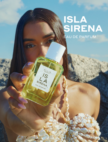 Woman with brown hair and a pink manicure wearing a white shell bikini top holds ISLA SIRENA Eau de Parfum out in front of her against a background of blue skies, white clouds, and gray rocks with white text that reads 'ISLA SIRENA Eau de Parfum'
