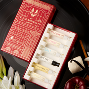 Red An Ellis Brooklyn Mystery Fragrance Discovery Set box opened to show inside and Apple Love 1.5ml Eau De Parfum Vial, Florist 1.5ml Eau De Parfum Vial, Salt 1.5ml Eau De Parfum Vial, Myth 1.5ml Eau De Parfum Vial, Vanilla Milk 1.5ml Eau De Parfum Vial and Sweet 1.5ml Eau De Parfum Vial on black vinyl record with vanilla beans, marshmallows, apple, sandalwood and white flowers, on wooden table.