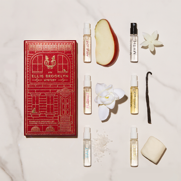 Red An Ellis Brooklyn Mystery Fragrance Discovery Set box on white marble table with Apple Love 1.5ml Eau De Parfum Vial next to apple slice, Myth 1.5ml Eau De Parfum Vial next to small white flower, Florist 1.5ml Eau De Parfum Vial next to white flower, Vanilla Milk 1.5ml Eau De Parfum Vial next to vanilla bean, Salt 1.5ml Eau De Parfum Vial next to sea salt, and Sweet 1.5ml Eau De Parfum Vial next to marshmallow.