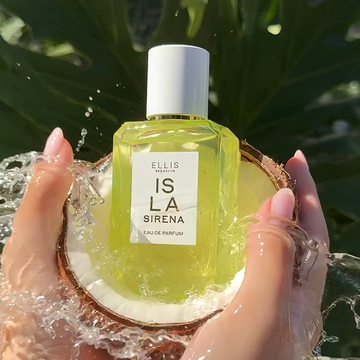 ISLA SIRENA Eau de Parfum 50ml sits in half of a coconut held in two hands with a pink manicure with a splash of water hitting it all against a background of green leaves