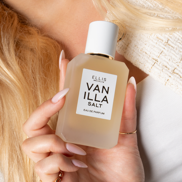 Woman with blonde hair and a pale pink manicure wearing a white tank top, a white and gold tweed jacket, and gold rings holds VANILLA SALT Eau de Parfum 50ml against her chest