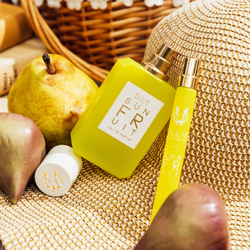 Sun Fruit 50ml Eau De Parfum with cap removed and Sun Fruit 10ml Eau De Parfum Travel Spray leaning on tan straw hat next to pears, books, and a wicker basket. 