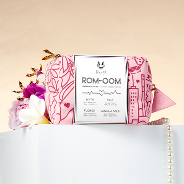 Red and pink pattered Rom-Com Fragrance Gift Set bag on white table with string of pearls, pink flower, purple flower, white flower, and yellow flower, on light orange backdrop. 