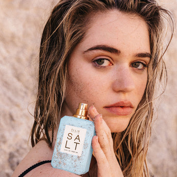 Model with damp blonde hair wearing black tank top, with sand on face and hand holding Salt 50ml Eau De Parfum with cap removed and sand on bottle, on shoulder.  