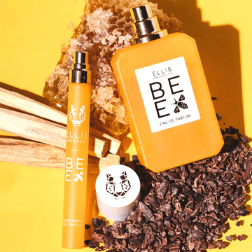 Bee 10ml Eau De Parfum travel Spray with cap removed and Bee 50ml Eau De Parfum with cap removed resting on piece of honey comb, pieces on wood, and cocoa absolute, on yellow background.