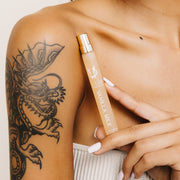 Model in strapless beige top with dragon tattoo and white nail polish holding Vanilla Milk 10ml Eau De Parfum Travel Spray below collarbone on white background.