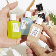 Model holding  Myth 7.5ml Eau De Parfum Mini Dabber, Salt 7.5ml Eau De Parfum Mini Dabber, Sun Fruit 7.5ml Eau De Parfum Mini Dabber, and Vanilla MIlk 7.5ml Eau De Parfum Mini Dabber in both hands over white flowers, sandalwood, green leaves, and cedarwood, on white background.