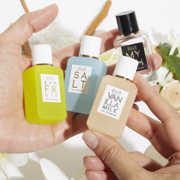 Model holding  Myth 7.5ml Eau De Parfum Mini Dabber, Salt 7.5ml Eau De Parfum Mini Dabber, Sun Fruit 7.5ml Eau De Parfum Mini Dabber, and Vanilla MIlk 7.5ml Eau De Parfum Mini Dabber in both hands over white flowers, sandalwood, green leaves, and cedarwood, on white background.