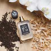 Myth 50ml Eau De Parfum with cap removed on wooden table next to white orchid flowers, amberette seeds, and white cedarwood shavings. 