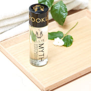 Myth Spectacular Scented Body Oil 95ml and white flower on wooden board, next to folded white towels and green leaves, on white table.  