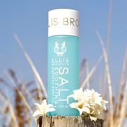 Salt Soothing Scented Body Oil 95ml next to white flowers on piece of wood with beach dunes and sky in background. 