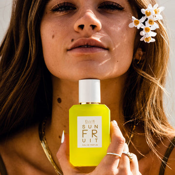 Model with brown hair and white nail polish wearing gold necklaces, earrings, and ring smiling white holding Sun Fruit 10ml Eau De Parfum Travel Spray near chin on white background. 