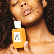 Model with brown curly hair holding Bee 50ml Eau De Parfum against jaw with honey dripping down cheek, jaw and lips, onto hand. 