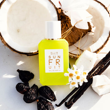 Sun Fruit 50ml Eau De Parfum leaning on opened coconut next to white flowers, vanilla beans, coconut pieces, and dried figs, on white background.